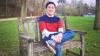 Mark, a male PhD candidate in his 20s sitting on a bench at the Downing College Paddock. He is wearing a pair of blue jeans, longsleeved polo and a colour-block sweater in dark blue, red and white. 